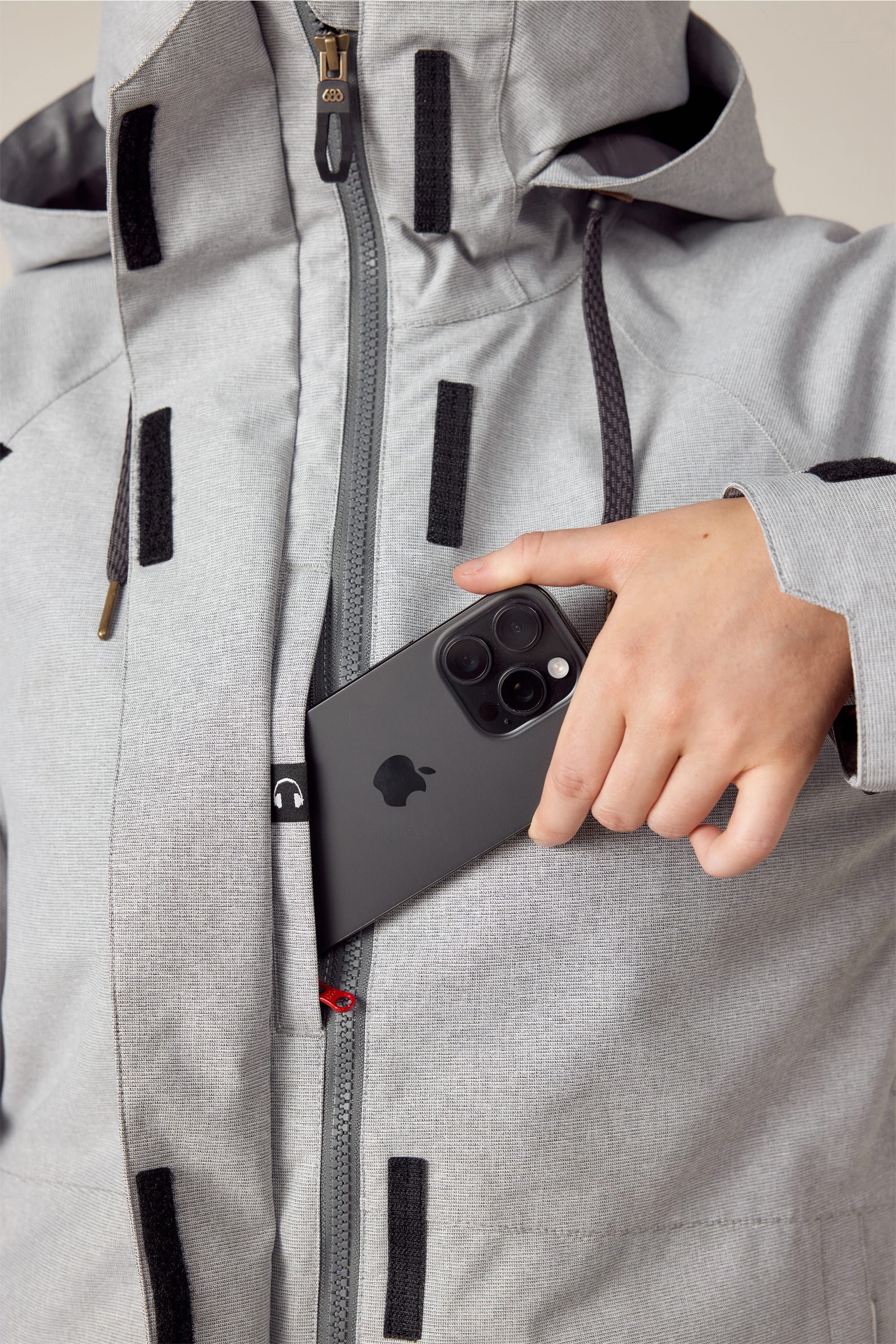 A person wearing the 686 Women's SMARTY® 3-In-1 Spellbound Jacket in light gray puts a black smartphone with three rear cameras into its zippered chest pocket.