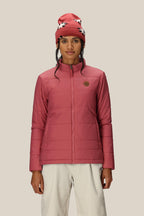 A woman wearing the 686 Women's SMARTY® 3-In-1 Spellbound Jacket in pink, white pants, and a red beanie with white and black patterns stands facing forward against a beige background.