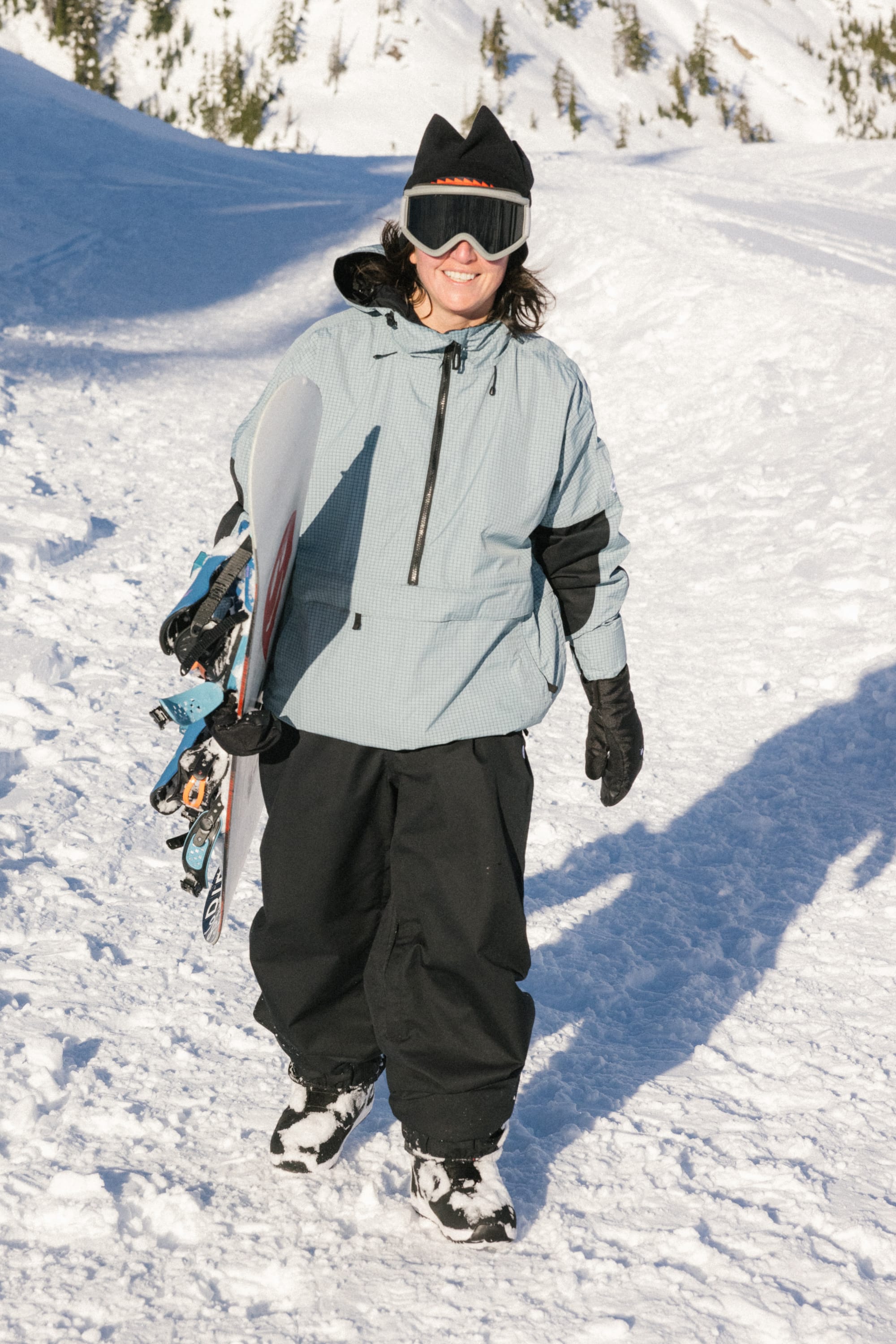 A woman wearing the 686 Women's Outline™ Shell Anorak smiles, holding a snowboard while walking on a snowy slope with trees, dressed in black goggles and gloves.