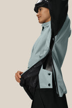 A woman in a light blue checkered jacket unzips the 686 Women's Outline™ Shell Anorak, revealing its inner lining. She is viewed from the side with a slight smile, all set against a plain beige background.