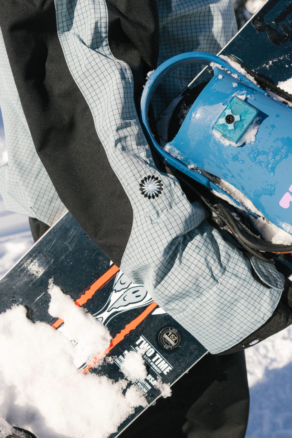 Wearing the 686 Women's Outline™ Shell Anorak, a person holds a snowboard with blue bindings in the snow. Snow covers part of the board, and the person's arm and hand are visible.