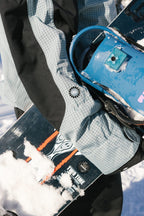 Wearing the 686 Women's Outline™ Shell Anorak, a person holds a snowboard with blue bindings in the snow. Snow covers part of the board, and the person's arm and hand are visible.