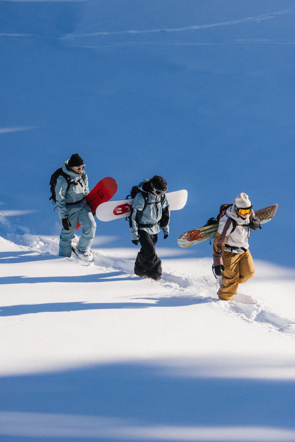 Three snowboarders, one wearing the 686 Women's Outline™ Shell Anorak, trek single file through deep, untouched snow carrying snowboards as they ascend a sunlit slope.