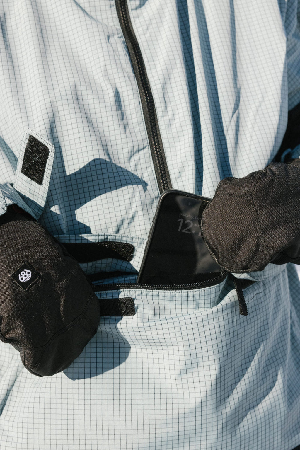 A person wearing black gloves and a 686 Women's Outline™ Shell Anorak places a smartphone into the jacket’s zippered front pocket.