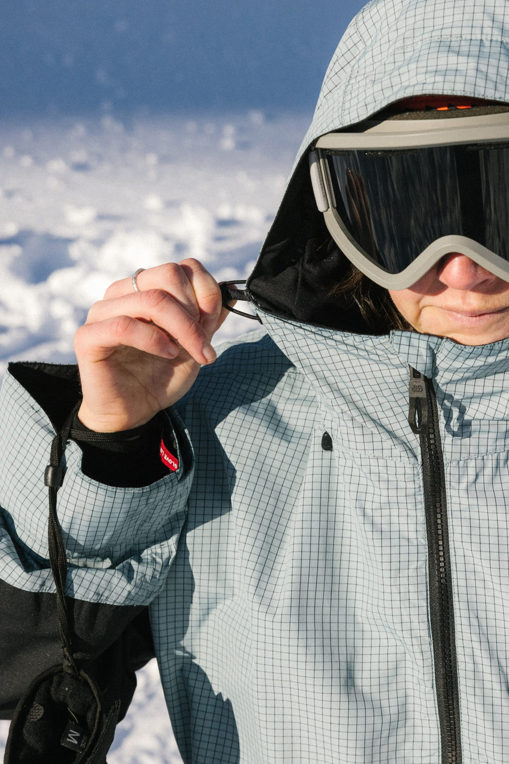 A woman wearing the 686 Women's Outline™ Shell Anorak and ski goggles adjusts her hood outdoors in bright, snowy conditions.