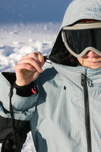 A woman wearing the 686 Women's Outline™ Shell Anorak and ski goggles adjusts her hood outdoors in bright, snowy conditions.