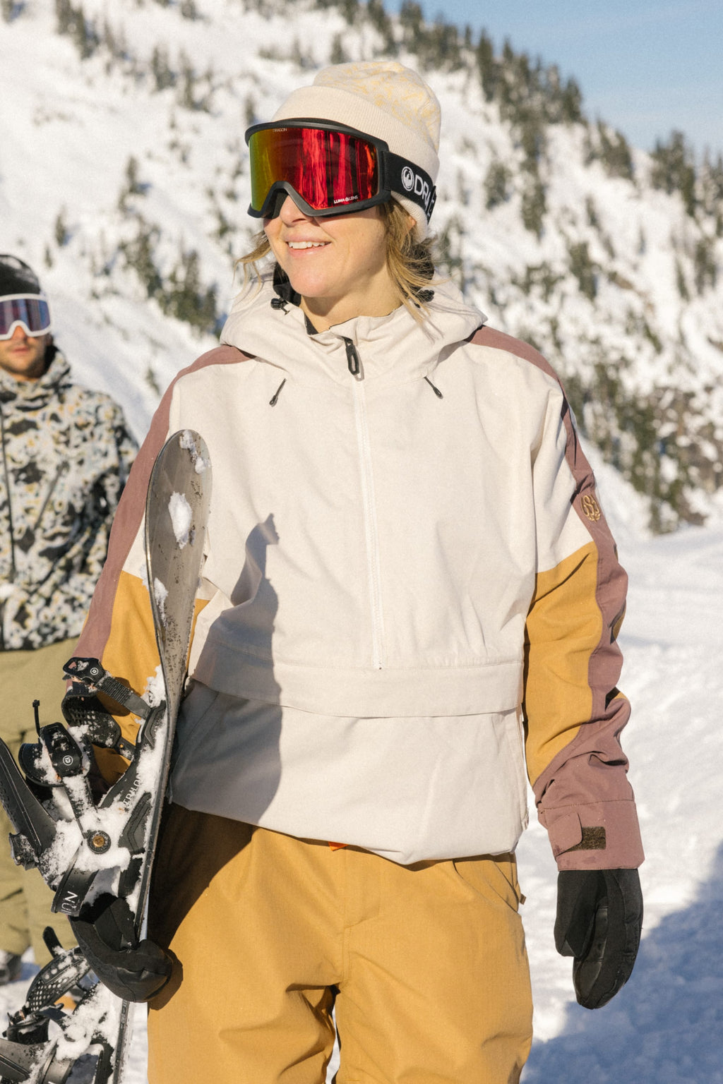A woman wearing the 686 Women's Outline™ Shell Anorak smiles holding a snowboard in snowy mountains, with another person visible in the background.