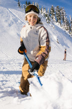 A woman smiles while shoveling snow on a sunny mountain slope, wearing the 686 Women's Outline™ Shell Anorak. Snow-covered trees and another person appear in the background.