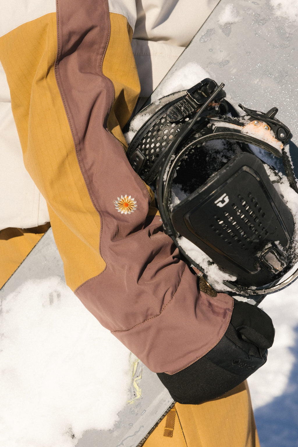 Wearing the 686 Women's Outline™ Shell Anorak in brown and yellow, a person rests their gloved hand on a snow-dusted snowboard. The snowboard binding and an embroidered flower on the sleeve are visible.
