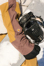 Wearing the 686 Women's Outline™ Shell Anorak in brown and yellow, a person rests their gloved hand on a snow-dusted snowboard. The snowboard binding and an embroidered flower on the sleeve are visible.