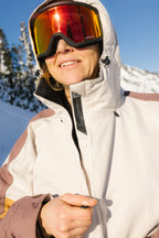A woman wearing the 686 Women's Outline™ Shell Anorak smiles outdoors on a sunny, snowy day with trees and mountains in the background, paired with reflective ski goggles.