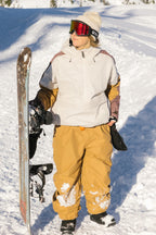 A person wearing the 686 Women's Outline™ Shell Anorak in white and tan, tan snow pants, and red ski goggles stands in the snow holding a snowboard, with a snowy landscape behind them.