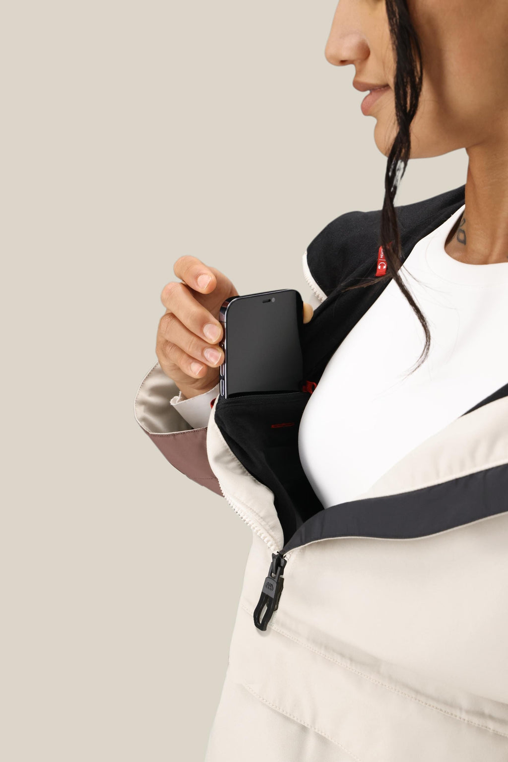 Wearing the 686 Women's Outline™ Shell Anorak, a person unzips the jacket partway to slip a smartphone into its inner chest pocket, revealing a white shirt beneath.