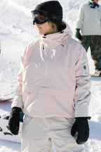 A woman wearing the 686 Women's Outline™ Shell Anorak stands on a snowy slope with white snow pants, black gloves, a black beanie, and ski goggles, surrounded by other snowboarders.