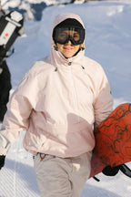 A woman in a light pink 686 Women's Outline™ Shell Anorak, white pants, black beanie, and ski goggles smiles while holding a red snowboard on a snowy mountain.