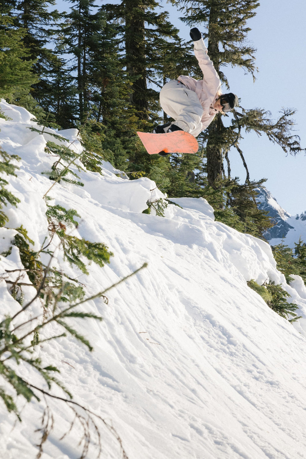 A snowboarder in the 686 Women's Outline™ Shell Anorak performs a midair trick above a snowy slope, evergreen trees in the background and a clear blue sky overhead.