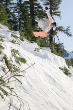 A snowboarder in the 686 Women's Outline™ Shell Anorak performs a midair trick above a snowy slope, evergreen trees in the background and a clear blue sky overhead.