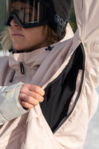 A person in the 686 Women's Outline™ Shell Anorak, a light pink waterproof jacket, unzips the underarm vent to show a black inner layer. Ski goggles and a snowy, tree-filled background complete the look.