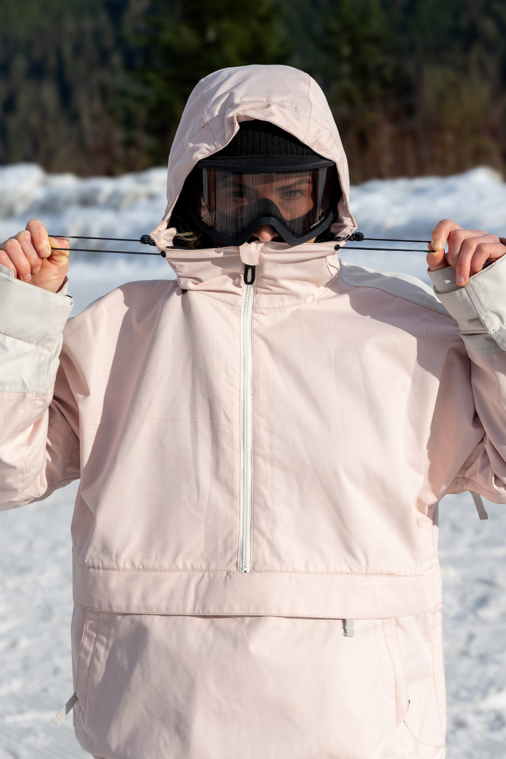 Wearing the 686 Women's Outline™ Shell Anorak, a black beanie, and ski goggles, a person stands outside in the snow, adjusting their hood drawstrings. Snow and trees are visible in the blurred background.