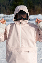 Wearing the 686 Women's Outline™ Shell Anorak, a black beanie, and ski goggles, a person stands outside in the snow, adjusting their hood drawstrings. Snow and trees are visible in the blurred background.