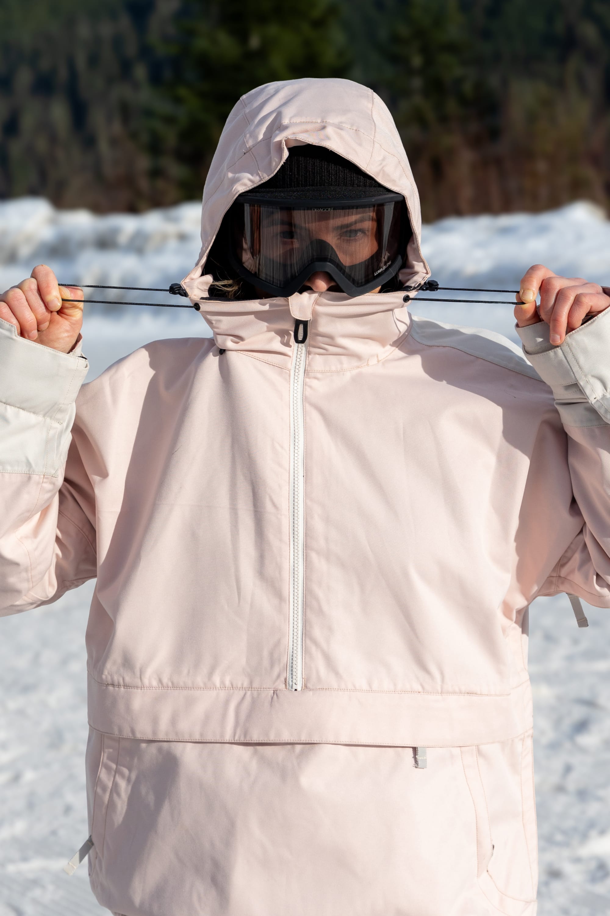 Wearing the 686 Women's Outline™ Shell Anorak, a black beanie, and ski goggles, a person stands outside in the snow, adjusting their hood drawstrings. Snow and trees are visible in the blurred background.