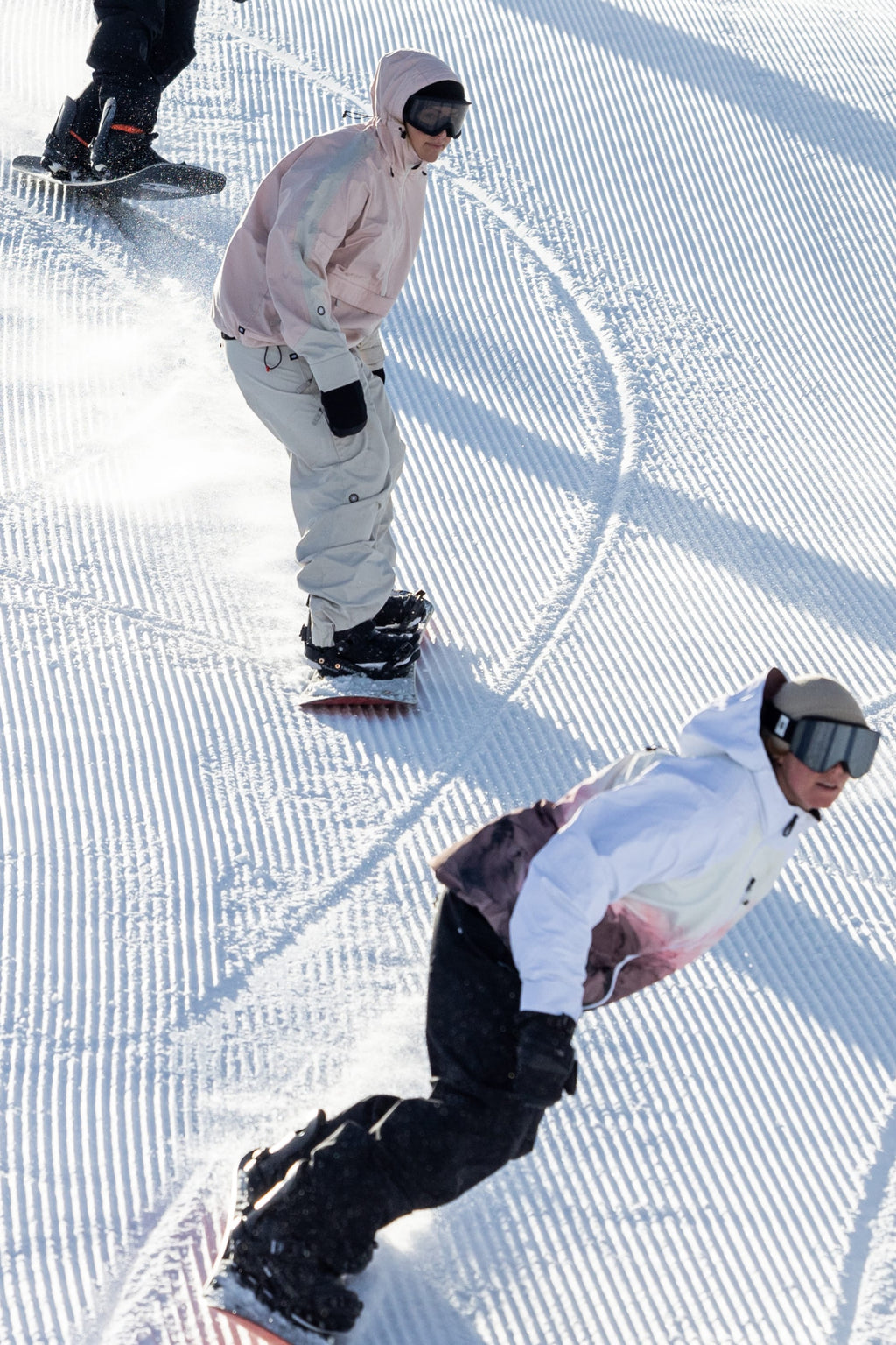 Snowboarders in 686 Women's Outline™ Shell Anorak jackets carve curved tracks down a groomed, snowy slope, sunlight casting shadows on the striped snow as they ride.