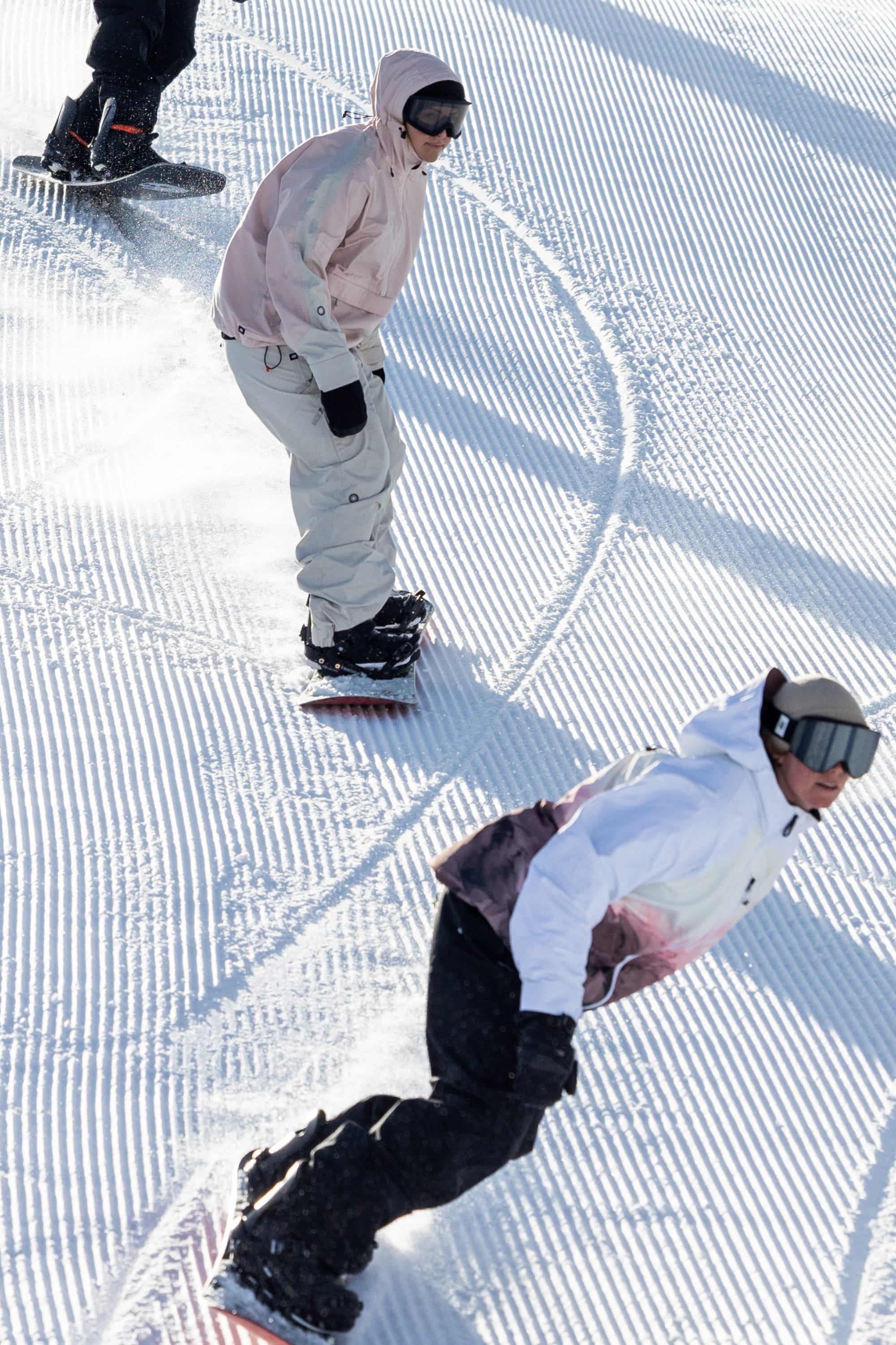 Snowboarders in 686 Women's Outline™ Shell Anorak jackets carve curved tracks down a groomed, snowy slope, sunlight casting shadows on the striped snow as they ride.