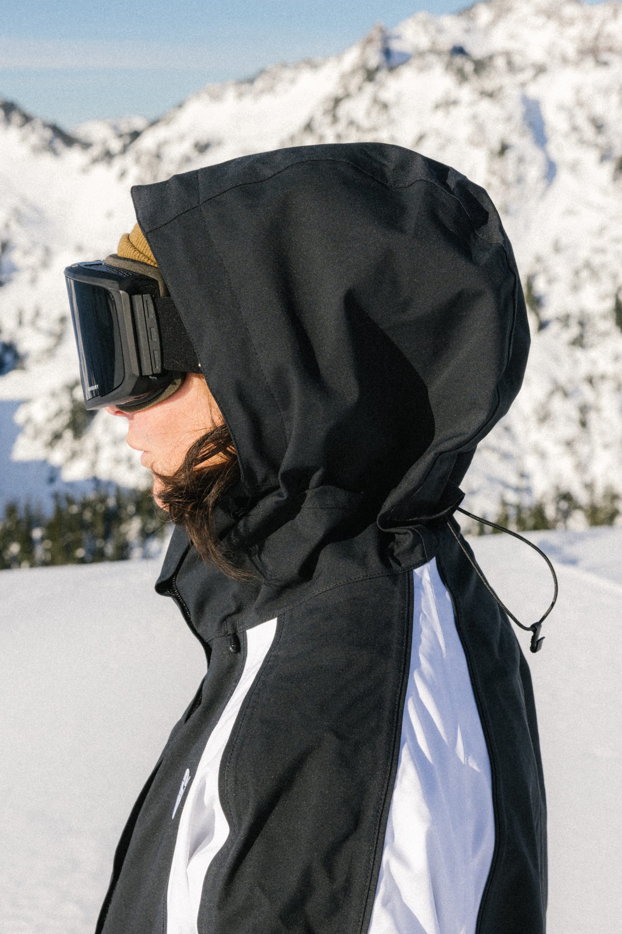 A person wearing the 686 Women's Hologram™ Shell Jacket, ski goggles, and a brown beanie stands in profile on a snowy mountain with sunlit peaks in the background.