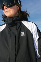 A person in the 686 Women's Hologram™ Shell Jacket and black ski goggles wears a brown hat outdoors under a blue sky.