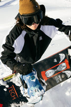 Wearing a 686 Women's Hologram™ Shell Jacket, a person stands in the snow with a brown beanie, black goggles, and holds a snowboard featuring colorful graphics and patches of snow.