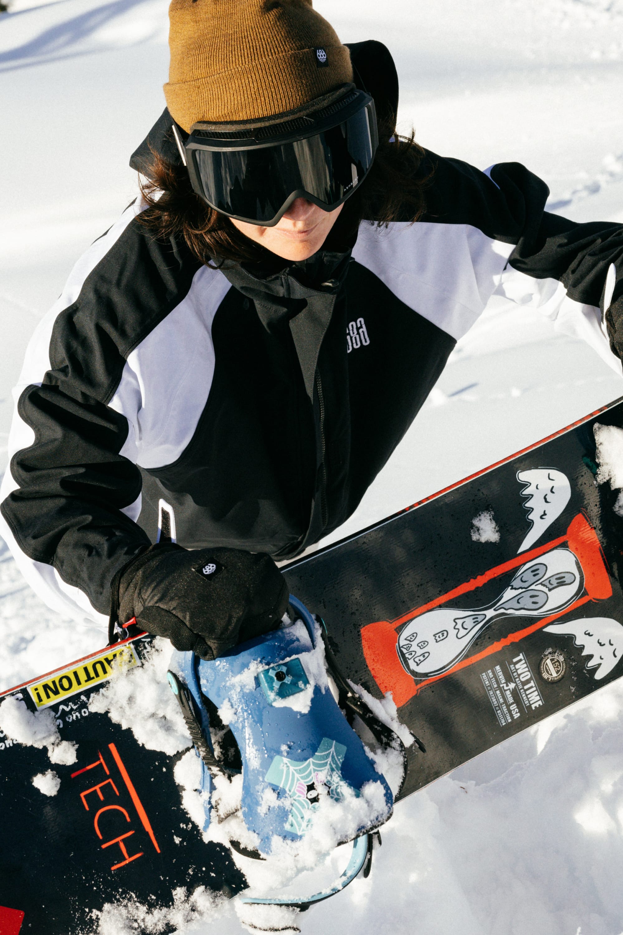 Wearing a 686 Women's Hologram™ Shell Jacket, a person stands in the snow with a brown beanie, black goggles, and holds a snowboard featuring colorful graphics and patches of snow.