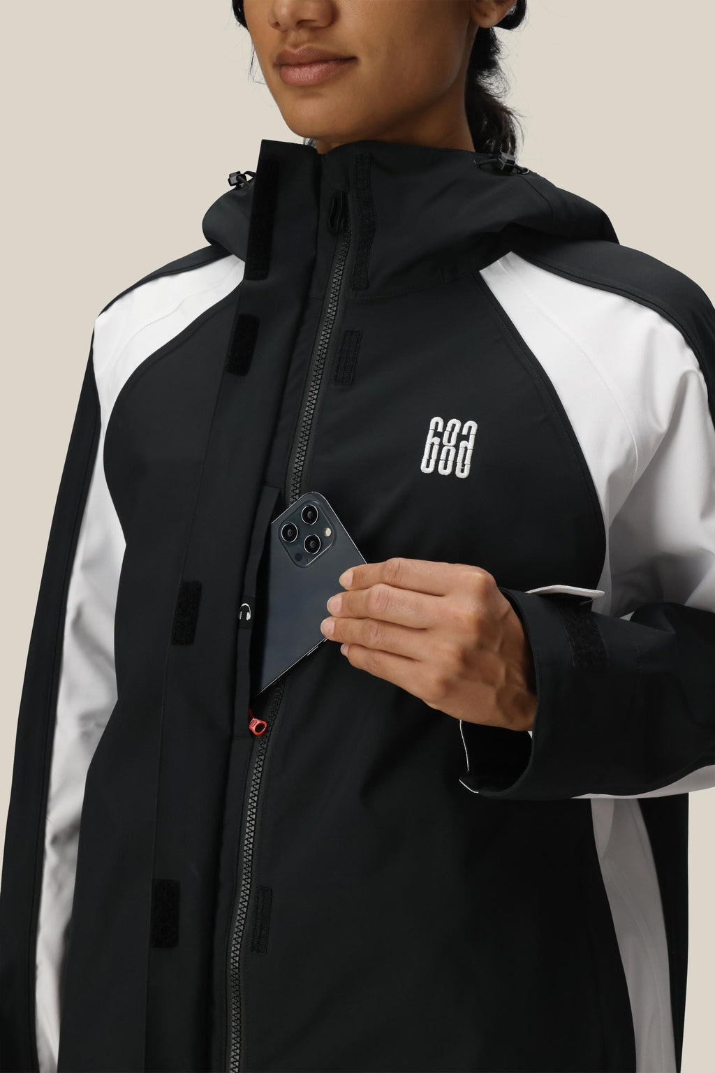 Wearing the 686 Women's Hologram™ Shell Jacket, a person slides a black smartphone into its zippered chest pocket. The black and white woven jacket features a 686 logo, and the person is set against a light beige background.
