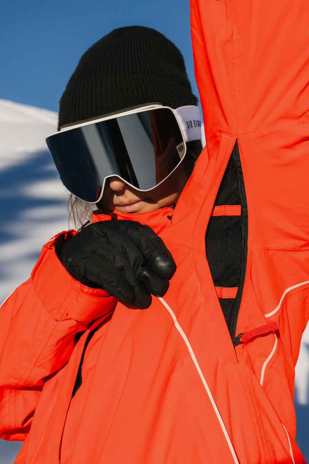 Wearing the 686 Women's Hologram™ Shell Jacket, a person in black gloves, a black beanie, and large ski goggles unzips their jacket under the arm, with snow and a blue sky in the background.