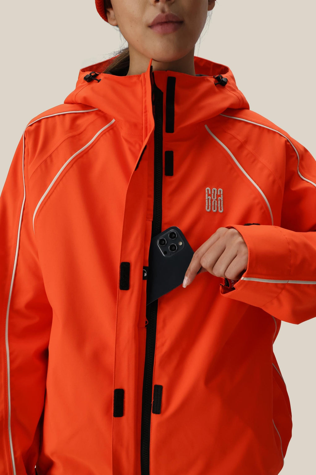A woman wearing the 686 Women's Hologram™ Shell Jacket in bright orange with white piping places a black smartphone into the chest pocket. The polyester jacket has a small white 686 logo on the left, set against a plain, light background.