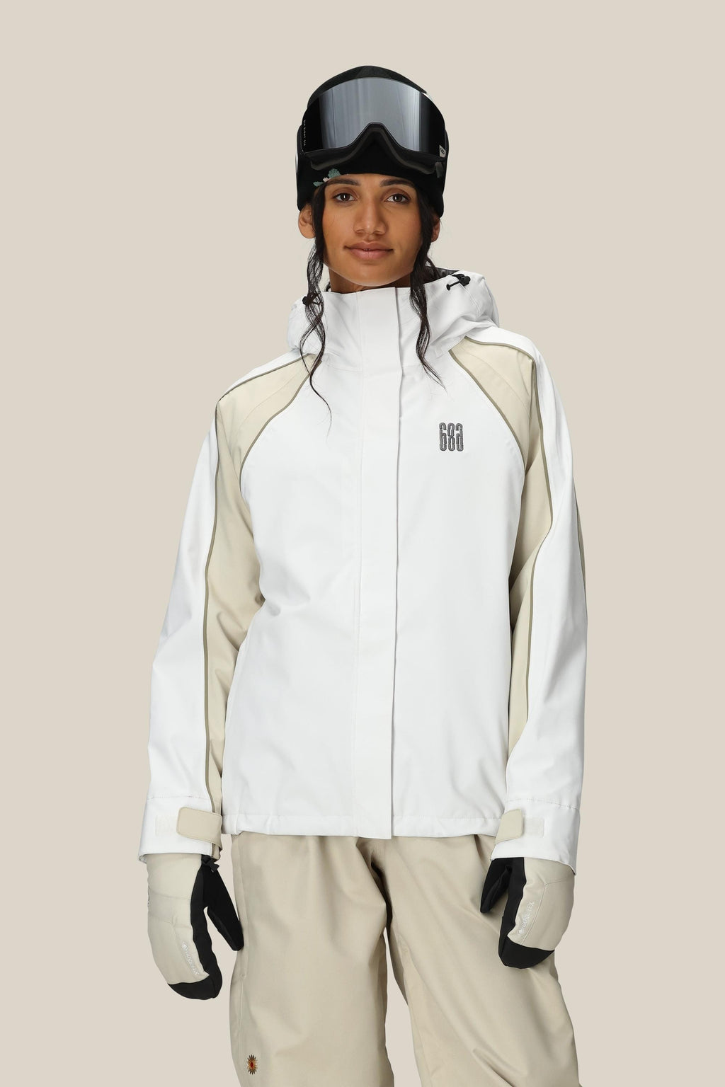 A woman wearing the 686 Women's Hologram™ Shell Jacket in white and beige stands facing forward against a plain light background, paired with beige ski pants, white gloves, and black ski goggles.
