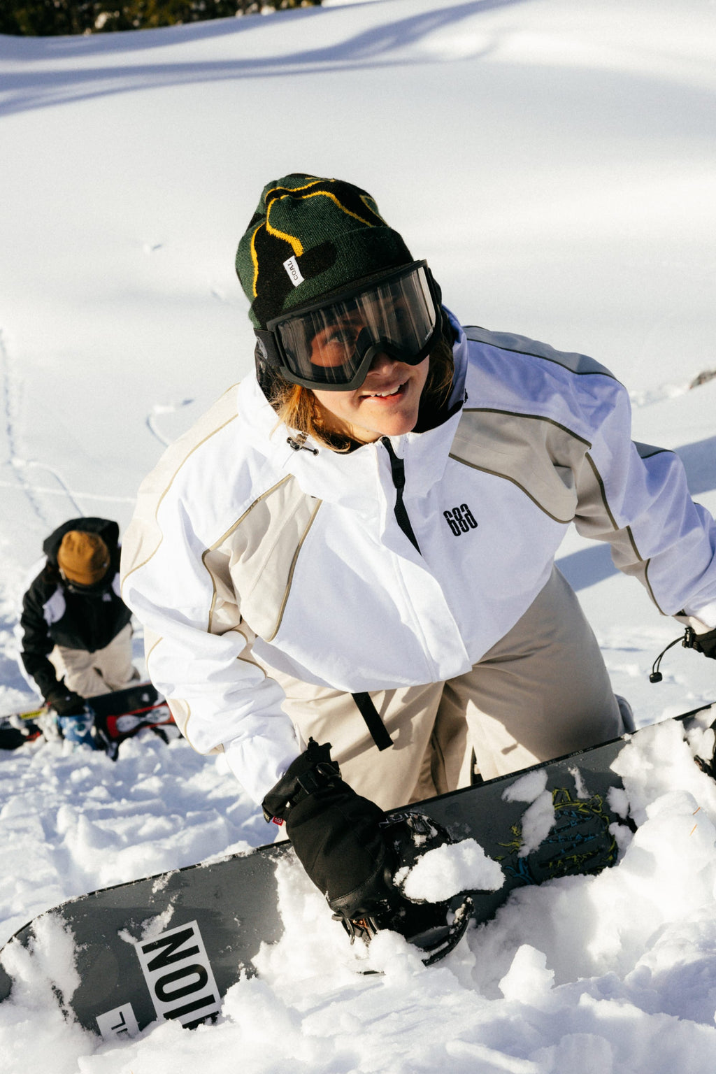 Two people in winter gear climb a snowy slope with snowboards. In front, someone wearing the 686 Women's Hologram™ Shell Jacket by 686, black gloves, goggles, and a green beanie smiles as they ascend through the snow-covered landscape.