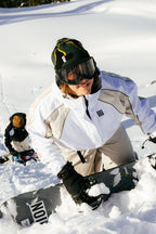 Two people in winter gear climb a snowy slope with snowboards. In front, someone wearing the 686 Women's Hologram™ Shell Jacket by 686, black gloves, goggles, and a green beanie smiles as they ascend through the snow-covered landscape.