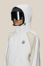 A person wearing the 686 Women's Hologram™ Shell Jacket in white with beige accents and large black ski goggles, standing against a plain light-colored background.