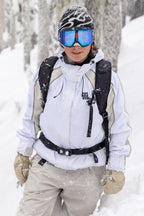 Wearing a 686 Women's Hologram™ Shell Jacket, beige pants, gloves, blue ski goggles, and a patterned beanie, a person stands in snowy woods with a black backpack as snow falls around them.