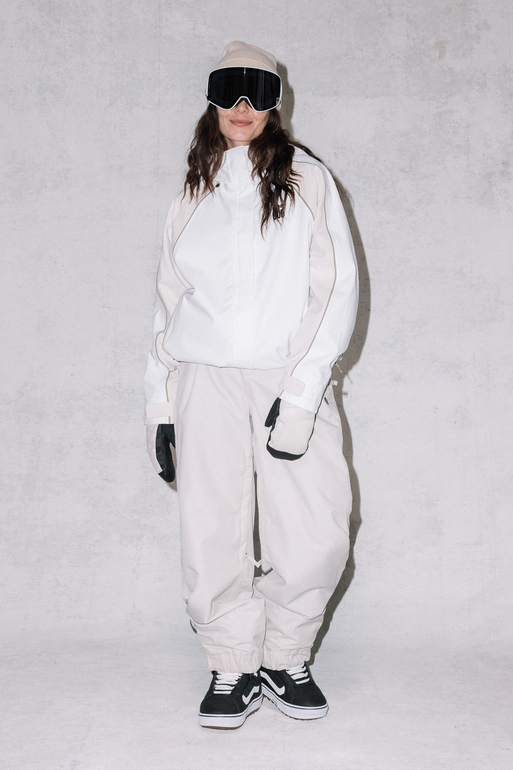 A woman wears the 686 Women's Hologram™ Shell Jacket by 686, styled as snow gear with black gloves, ski goggles, and sneakers, standing against a plain light gray background and smiling slightly.