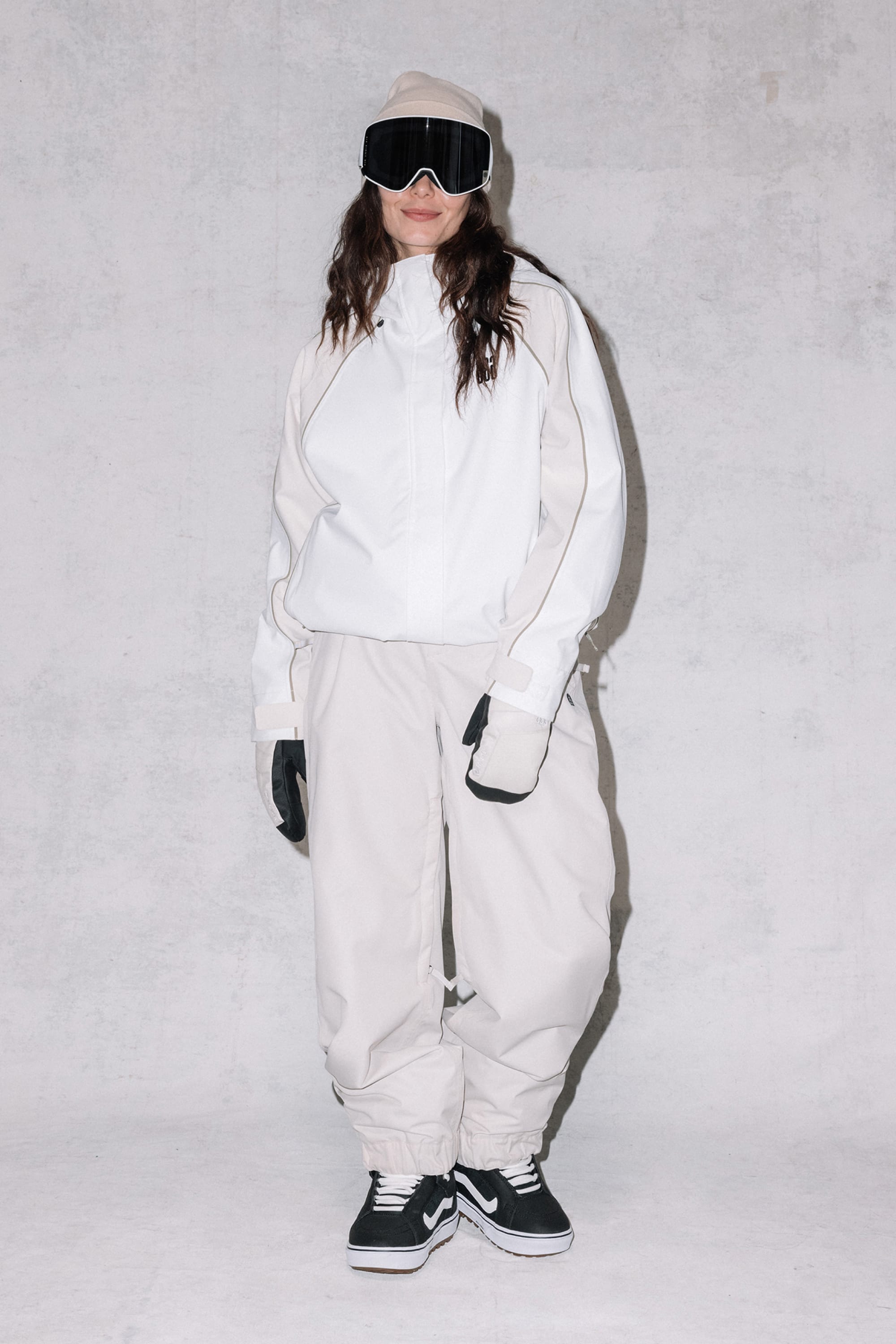 A woman wears the 686 Women's Hologram™ Shell Jacket by 686, styled as snow gear with black gloves, ski goggles, and sneakers, standing against a plain light gray background and smiling slightly.
