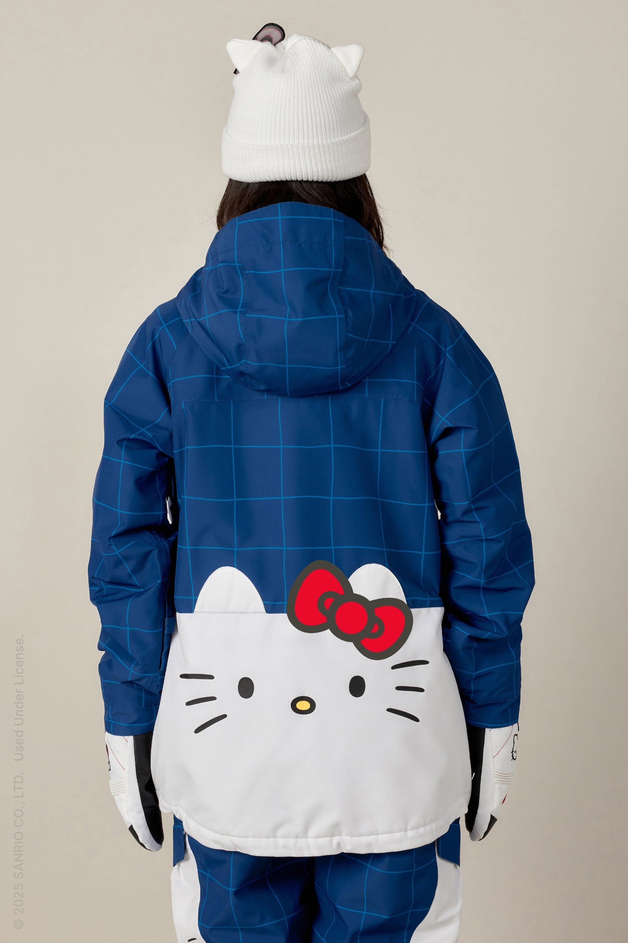 A person is seen from behind wearing the 686 Women's Athena™ Insulated Jacket by 686, which features a blue checkered design, a large Hello Kitty face with a red bow on the back, matching pants, white gloves, and a white beanie with Hello Kitty ears and bow.