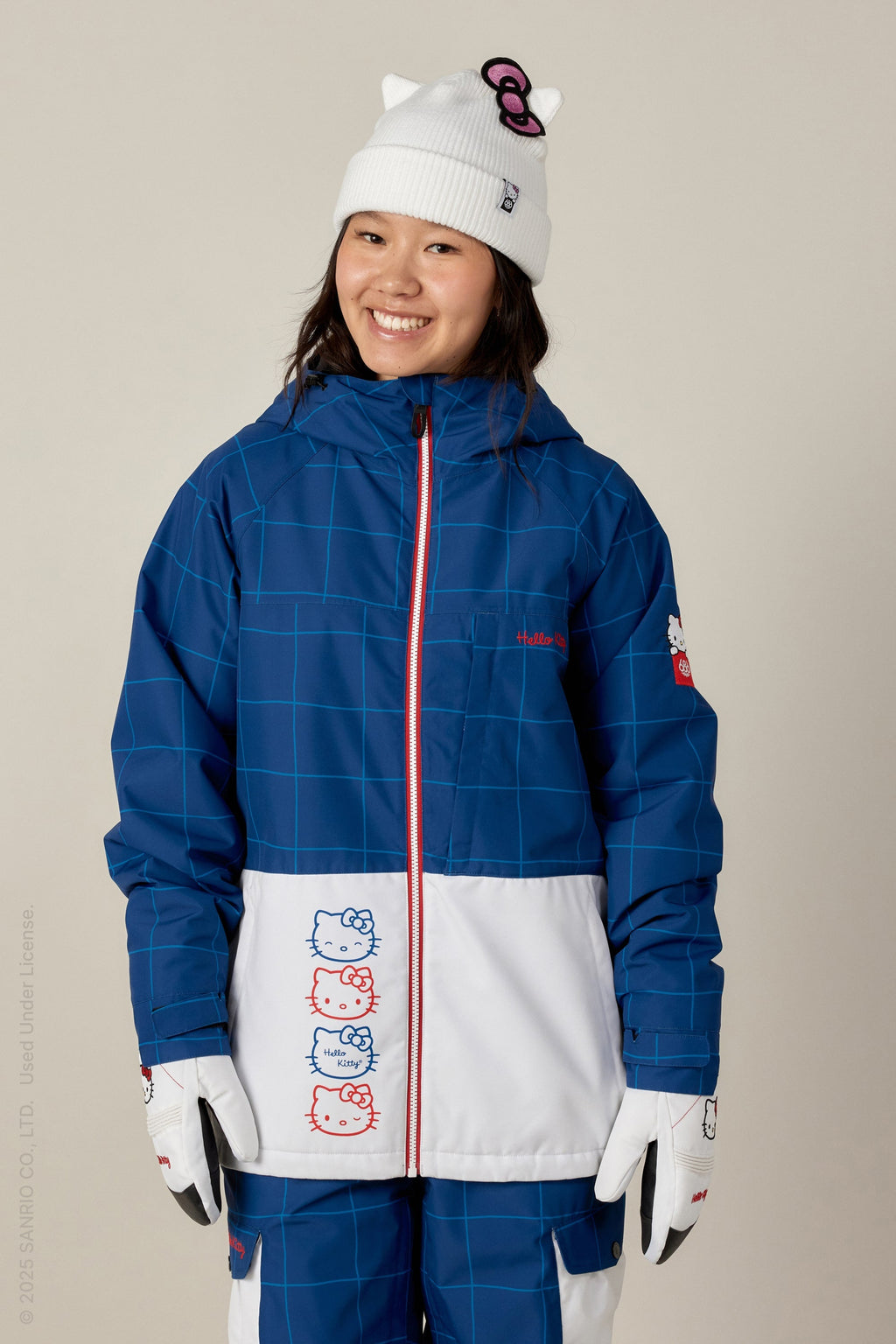 A smiling young woman wears the 686 Women's Athena™ Insulated Jacket by 686, paired with matching pants, white gloves, and a white beanie with cat ears and a bow.