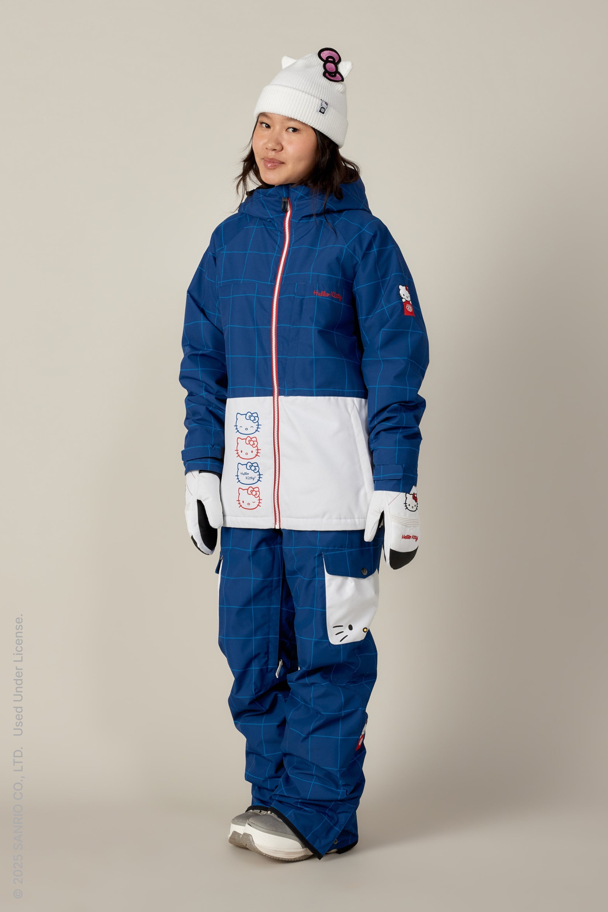 A person wearing the 686 Women's Athena™ Insulated Jacket in a blue and white Hello Kitty winter outfit, paired with matching pants, white gloves, and a white beanie featuring Hello Kitty’s face and a 3D red bow.