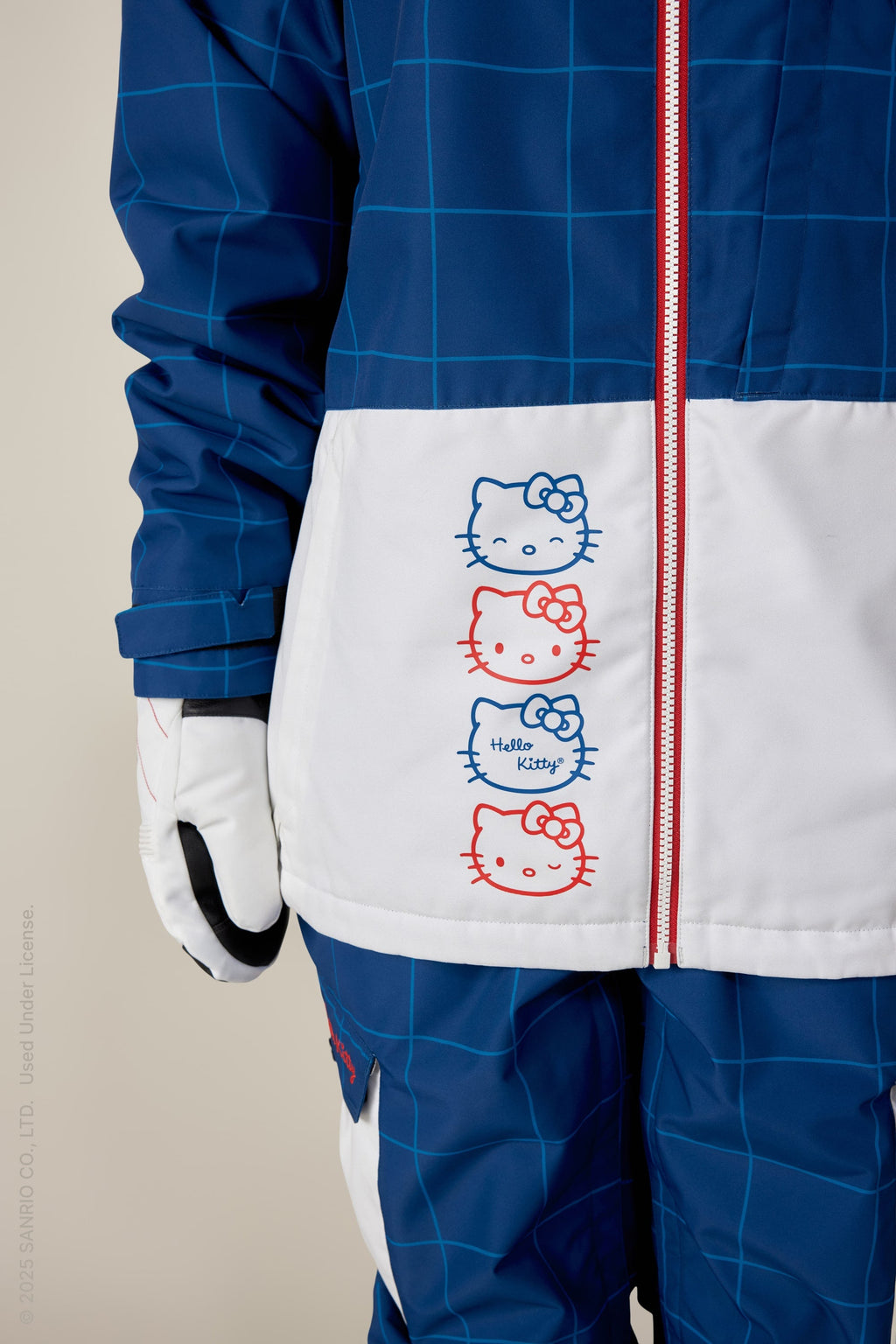 A person wears the 686 Women's Athena™ Insulated Jacket, a blue and white polyester waterproof jacket with red zippers. It features three vertically stacked Hello Kitty faces in red and blue outlines and the text Hello Kitty; only lower half and left arm are visible.