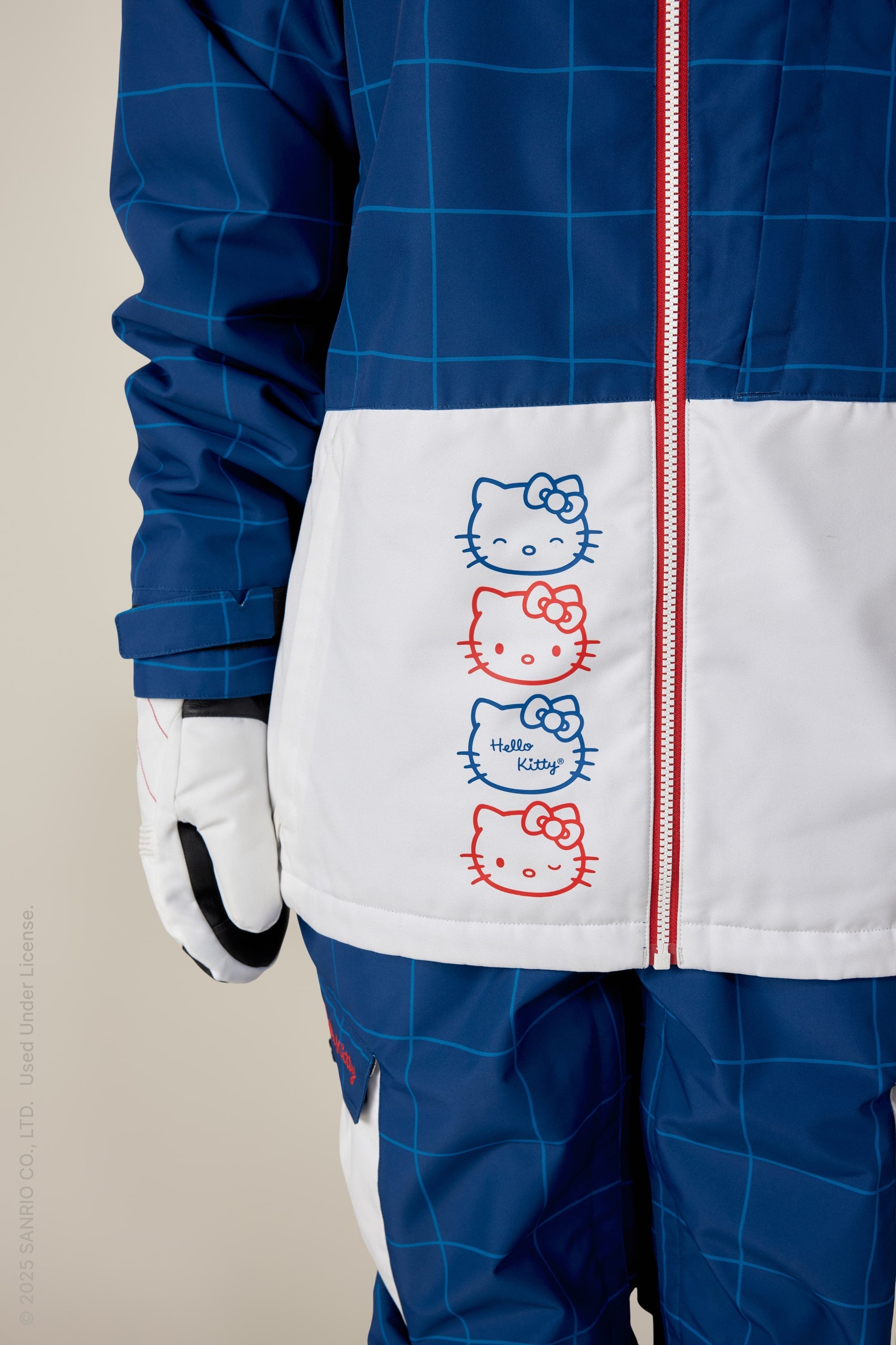 A person wears the 686 Women's Athena™ Insulated Jacket, a blue and white polyester waterproof jacket with red zippers. It features three vertically stacked Hello Kitty faces in red and blue outlines and the text Hello Kitty; only lower half and left arm are visible.