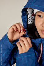 A woman wearing the 686 Women's Athena™ Insulated Jacket in blue check checks the hood’s drawstring. The inner hood displays a cartoonish black and red pattern, and only half of her face is visible under a white beanie.