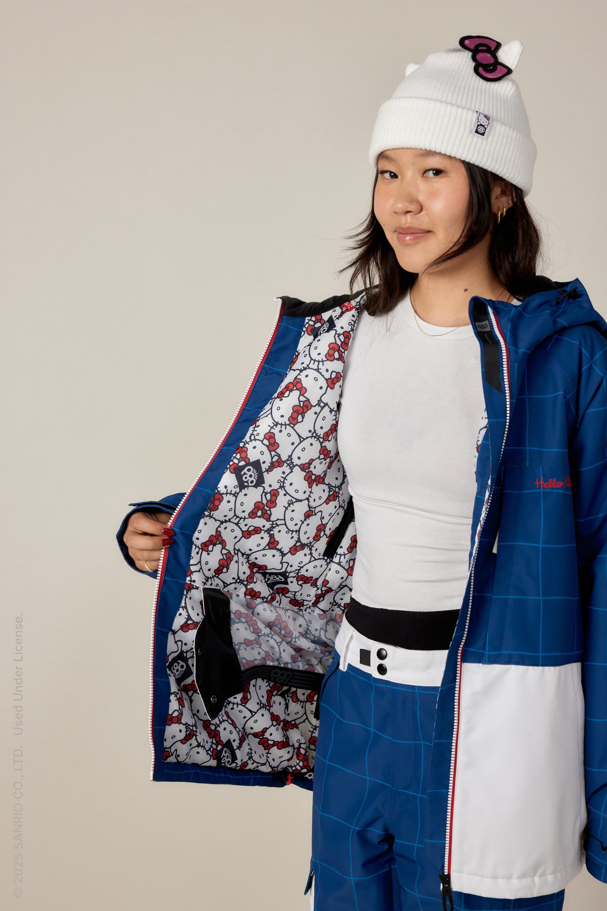 A young woman in a white Hello Kitty beanie and blue 686 Women's Athena™ Insulated Jacket opens her jacket to reveal a white Hello Kitty patterned lining. She wears blue snow pants and a white shirt, smiling slightly at the camera.
