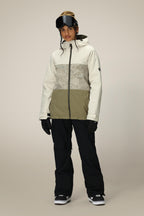 A person wears the 686 Women's Athena™ Insulated Jacket in beige and olive green, paired with black snow pants, gloves, black snow goggles on their head, and black snow boots, standing against a plain light background.
