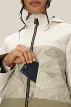 A woman in a light 686 Women's Athena™ Insulated Jacket puts a dark smartphone into the jacket’s front zip pocket. Her face is partly visible against a plain background.